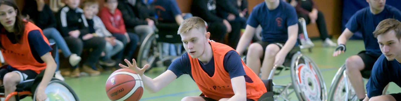 Jugendliche Jungs und Mädchen im Rollstuhl spielen Basketball. Ein Junge dribbelt den Ball. Im Hintergrund stehen viele Zuschauer.