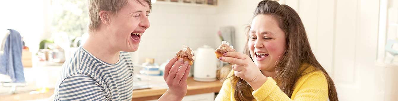 Ein junger Mann und eine junge Frau lachen herzlich und stoßen mit Muffins an. Sie sind in einer Küche und die Sonne scheint herein.