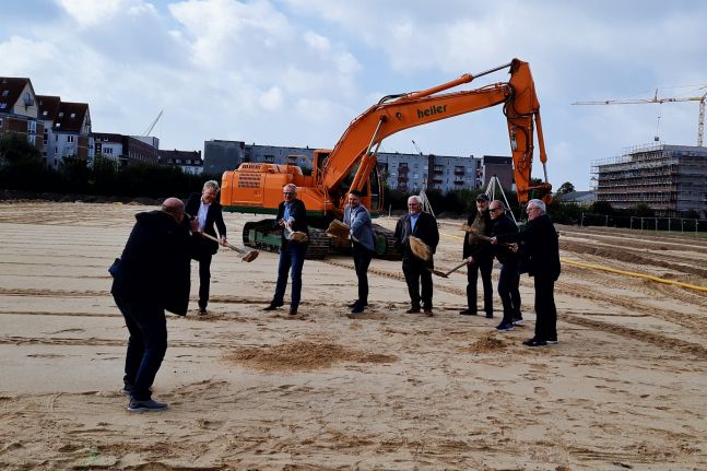 Auf einer großen Baustelle. Mehrere Männer mit Schaufeln in der Hand schaufeln symbolisch Sand. Im Hintergrund steht ein Bagger.