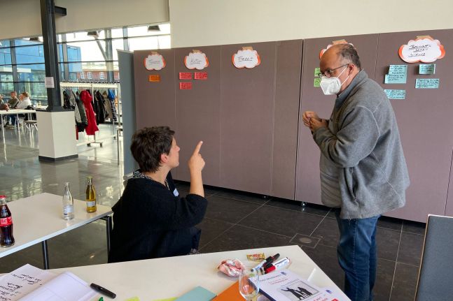 Ein Mann und eine Frau schauen auf die Arbeitsergebnisse einer Arbeitsgruppe, die auf Karten an eine Wand  geklebt wurden